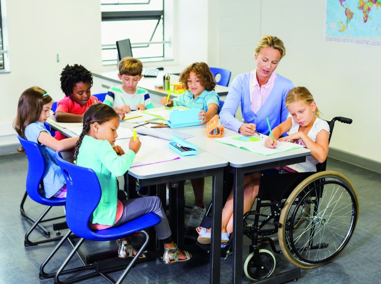 Photo : Dans une salle de classe, cinq enfants, dont un en fauteuil roulant, sont autour d’une grande table et écrivent dans leur cahier. Leur enseignante donne des conseils à l’enfant en situation de handicap.